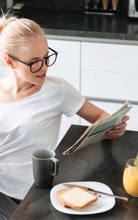 Serious focused lady reading newspaper while have breakfast Serious focused lady in eyeglasses reading newspaper while have breakfast in kitchen